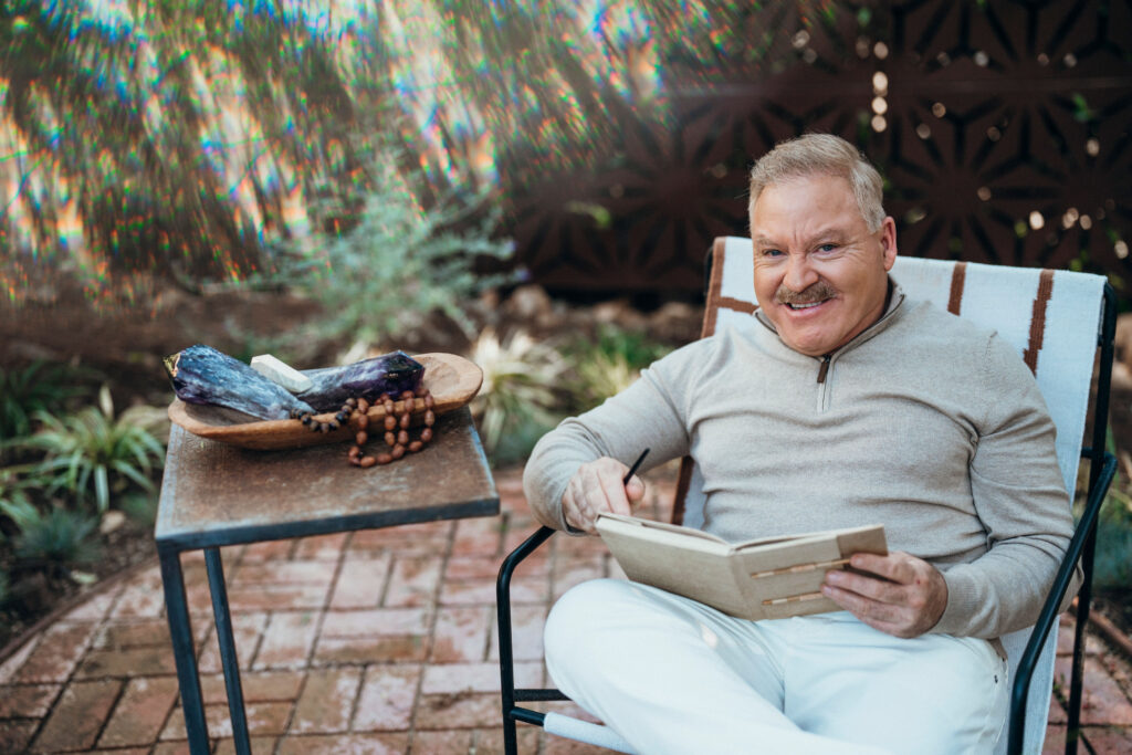 A man sits outdoors in a chair, holding a notebook and pen, smiling at the camera. A small table next to him has eggplant, grapes, and cheese on a wooden plate.