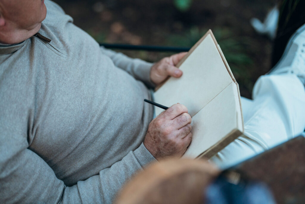 A person wearing a beige sweater is seated and holding an open blank notebook while writing with a pencil.