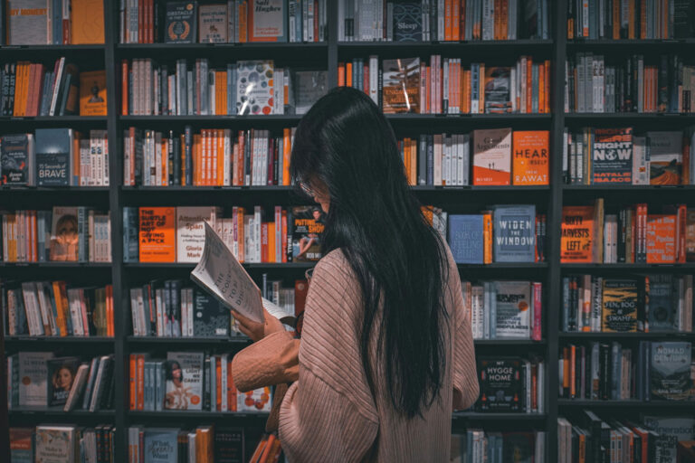 A person with long dark hair stands in front of a bookshelf, reading a book in a bookstore or library.