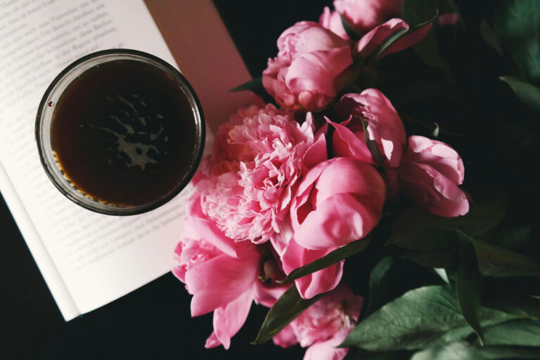 A glass of dark coffee rests on an open book beside a bouquet of pink peonies with green leaves.