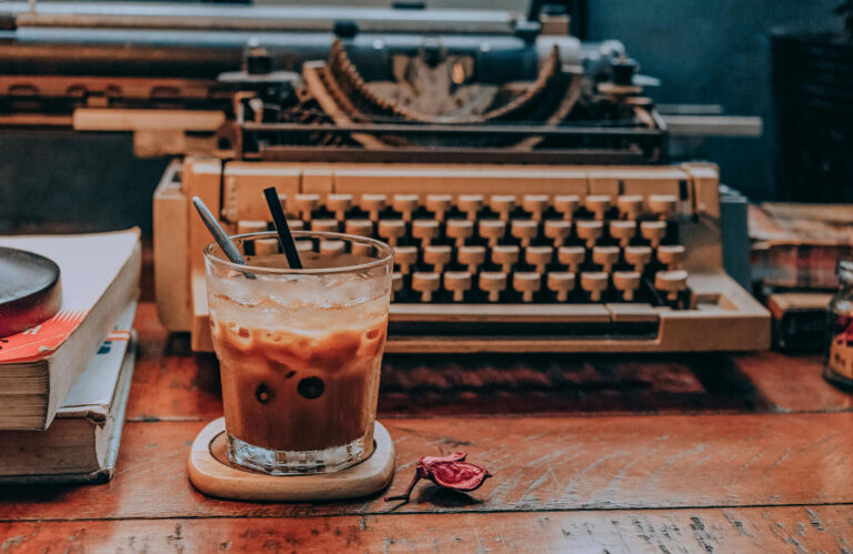 A glass of iced coffee with two straws sits on a coaster in front of a vintage typewriter on a wooden desk.