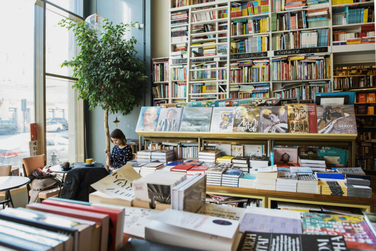A woman sits at a table with a drink, using her phone in a cozy bookstore filled with shelves and stacks of books by a large window.