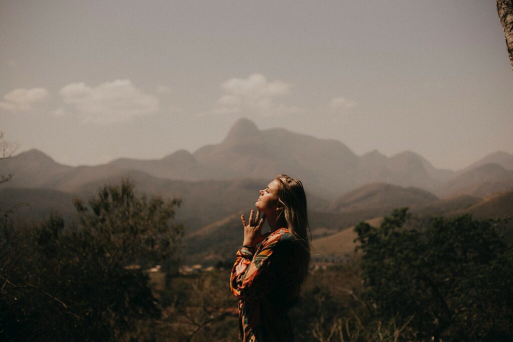 A woman stands outdoors with her eyes closed and hands clasped, facing sunlight, with mountains and trees in the background.