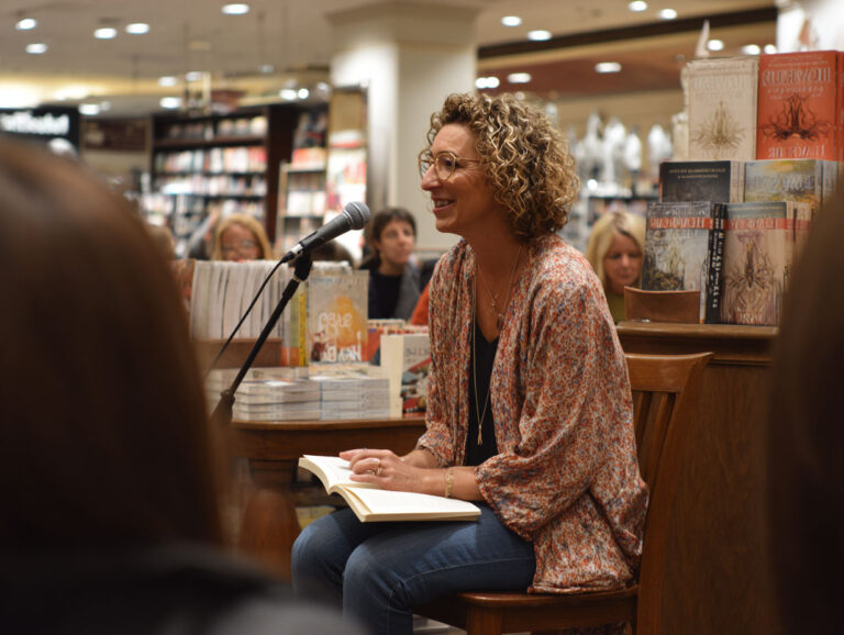A woman with curly hair sits on a chair, smiling and speaking into a microphone, holding an open book in a bookstore with shelves and people in the background.