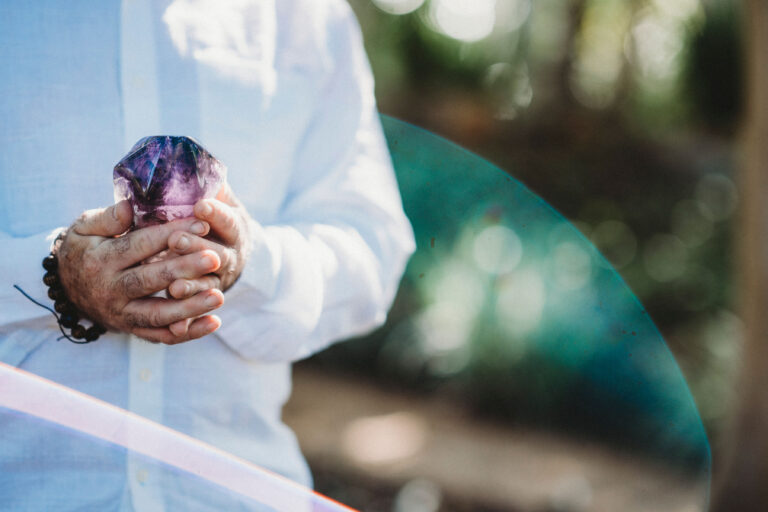 A person in a white shirt holds a large, faceted purple crystal with both hands; the background is outdoors and blurred.