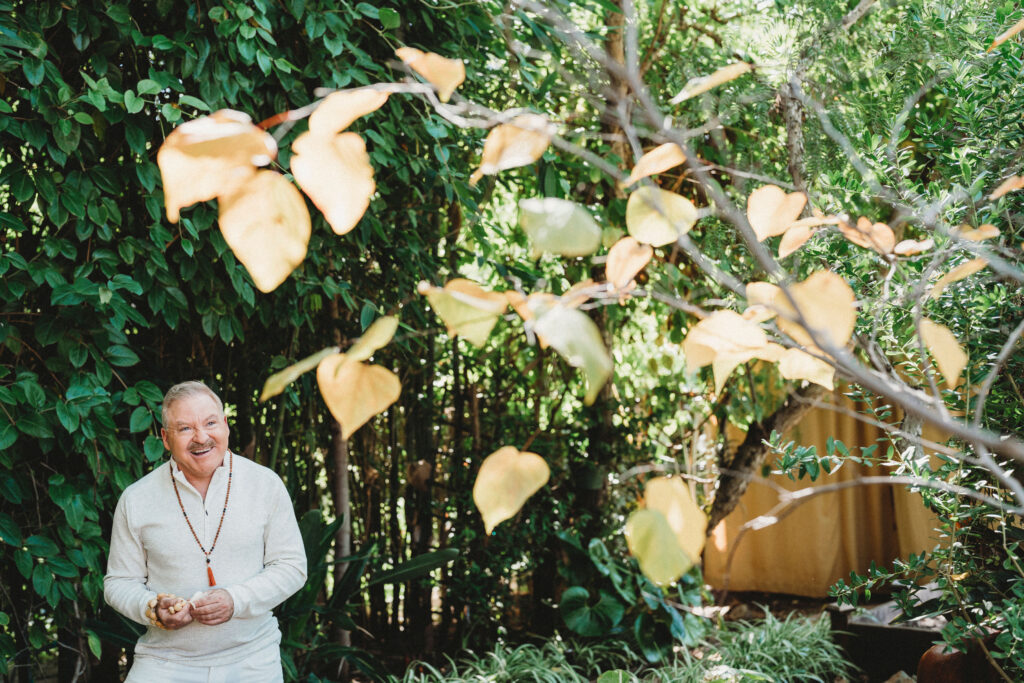 A man in a light-colored outfit stands smiling in a lush garden with green foliage and branches bearing yellow leaves.
