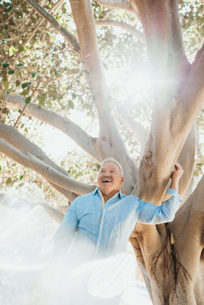 An older man in a light blue shirt stands smiling and holding a tree branch, with sunlight streaming through the branches behind him.