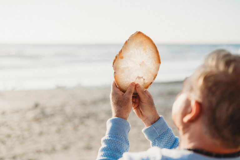 A person holds up a large slice of translucent mineral or crystal while standing on a sandy beach, facing the ocean.