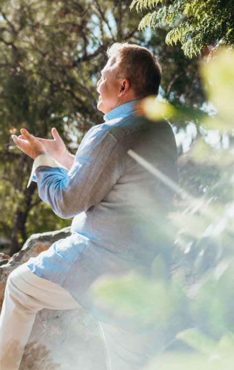 An older man sits outdoors on a rock with his hands raised, looking upward. Sunlight filters through trees in the background.