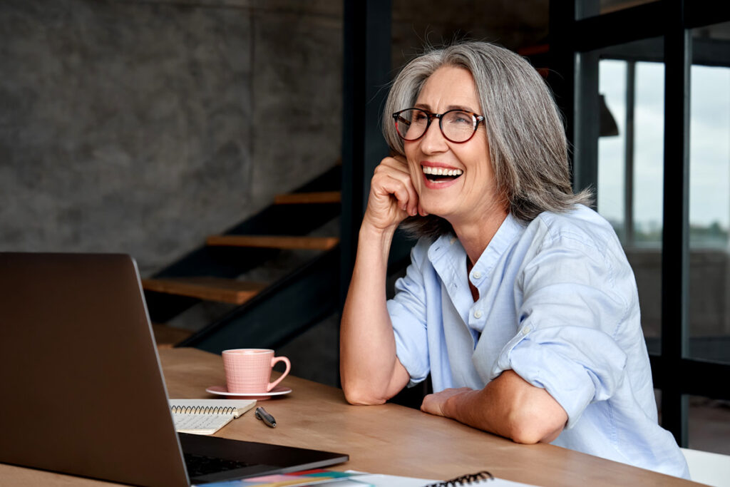 Smiling older woman with gray hair and glasses sits at a desk with a laptop, coffee cup, and notebooks, looking off-screen.