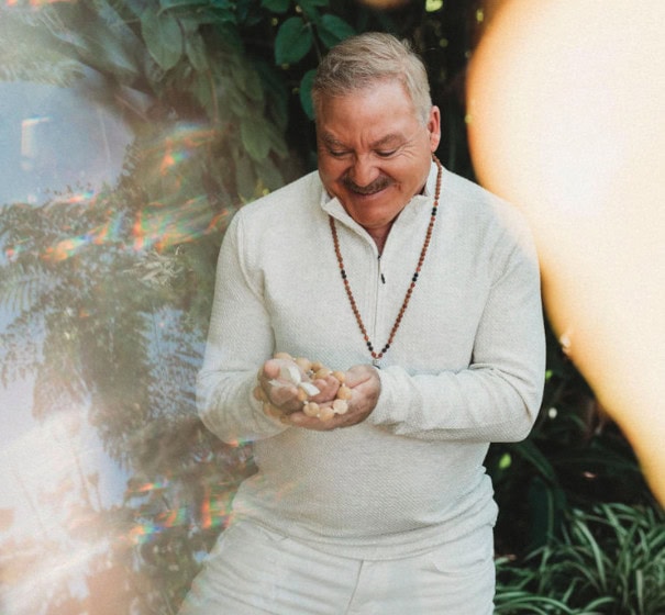 A man in a white outfit stands outside holding small stones in his hands, surrounded by greenery and sunlight.