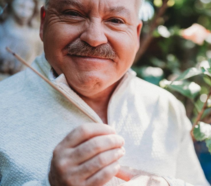 An older man with a mustache and light sweater smiles at the camera while holding a thin stick outdoors with greenery in the background.