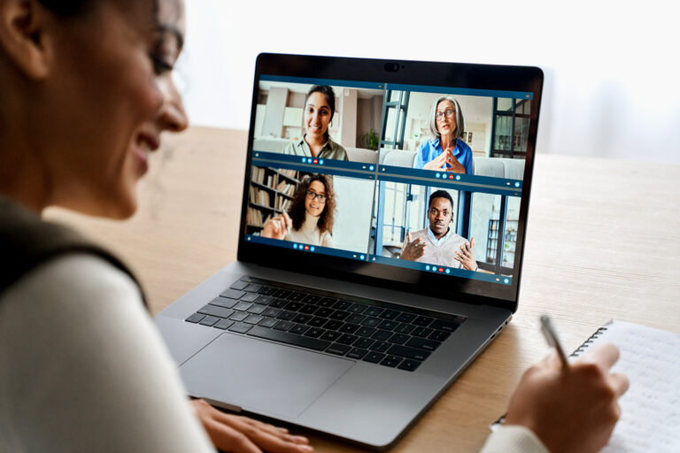 A person takes notes while participating in a video conference with four people displayed on a laptop screen.