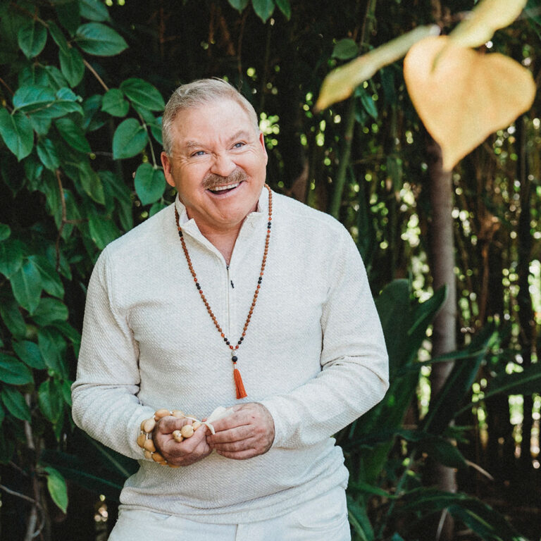 A man with a mustache wearing a light sweater, beaded necklace, and bracelets stands outdoors in front of green foliage, smiling and holding beads.