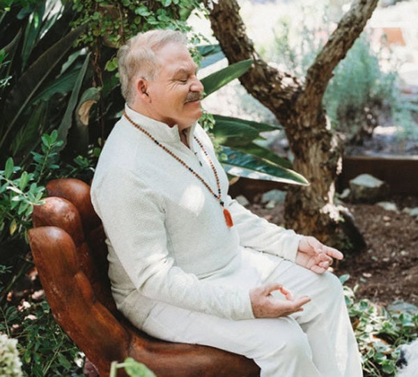 Older man in white clothing sits cross-legged on a hand-shaped chair outdoors, meditating with a string of prayer beads around his neck, surrounded by greenery.