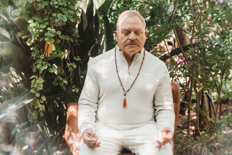 An older man in white clothing and a beaded necklace sits outdoors with eyes closed, meditating in a garden surrounded by green plants.