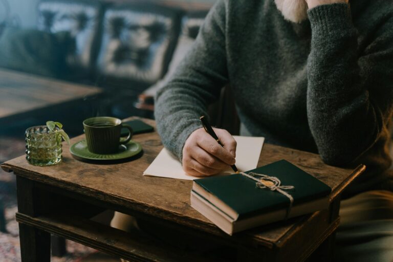 A person sits at a wooden table, writing on paper with a pen. A green teacup, saucer, and a tied notebook are also on the table. The background shows a sofa.