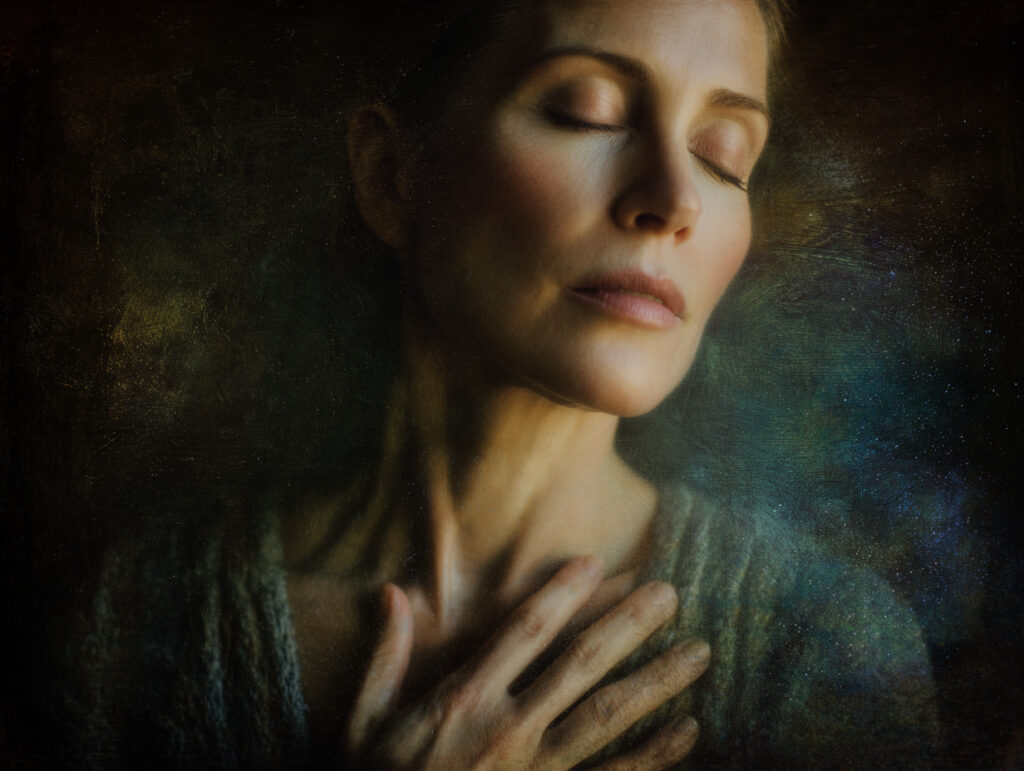 A woman with closed eyes and a peaceful expression rests her hand on her chest against a dark, textured background.