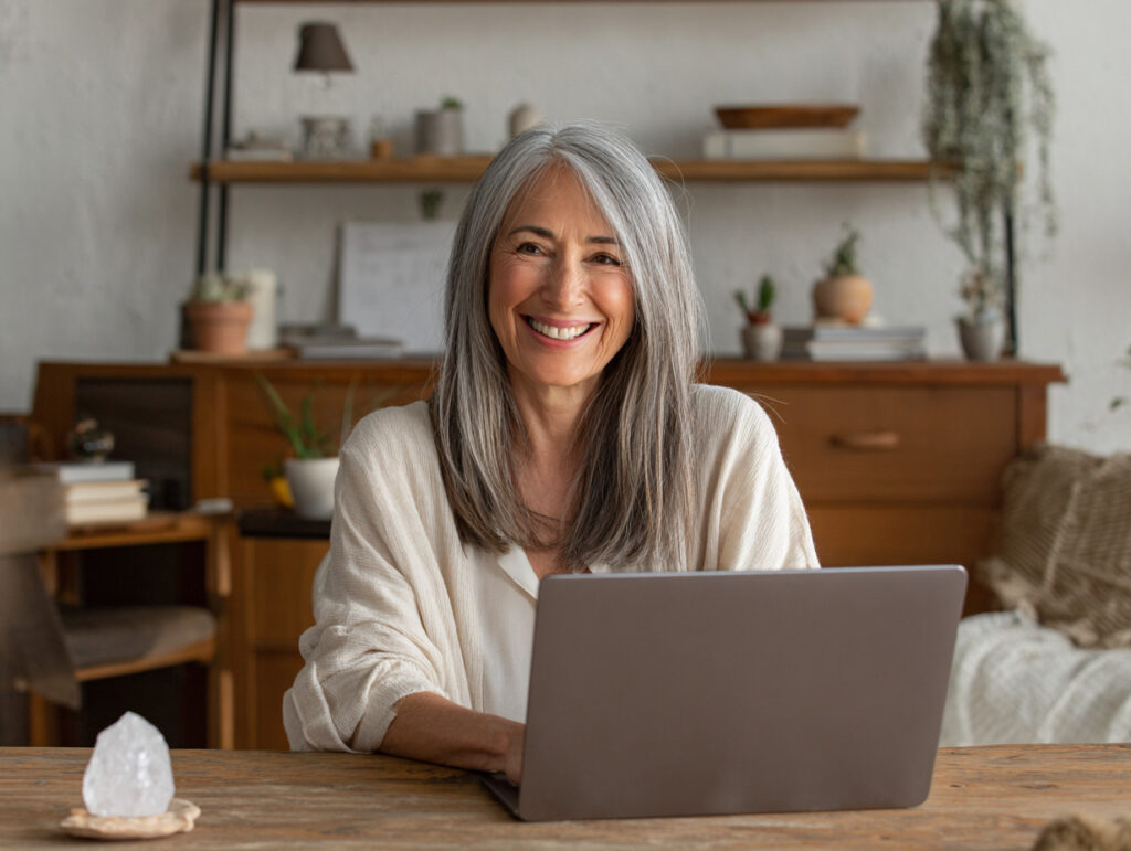A woman with long gray hair sits at a wooden table, smiling while using a laptop in a warmly decorated home office.
