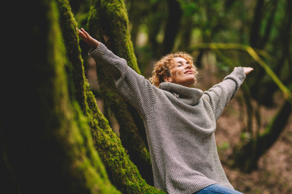 A person with curly hair wearing a gray sweater sits among mossy trees with arms outstretched and eyes closed, appearing relaxed in a forest setting.