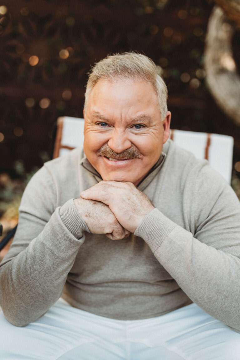 A middle-aged man with short gray hair and a mustache sits outdoors, smiling with his hands clasped under his chin.