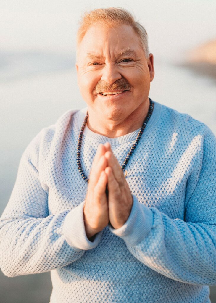Man with short light hair and mustache, wearing a light blue sweater and beaded necklace, stands outdoors with hands pressed together in a gesture of greeting or gratitude.